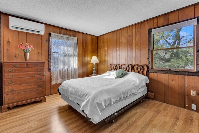 bedroom featuring a textured ceiling, wooden walls, light hardwood / wood-style floors, and a wall mounted air conditioner