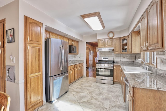 kitchen with backsplash, light stone countertops, under cabinet range hood, appliances with stainless steel finishes, and a sink