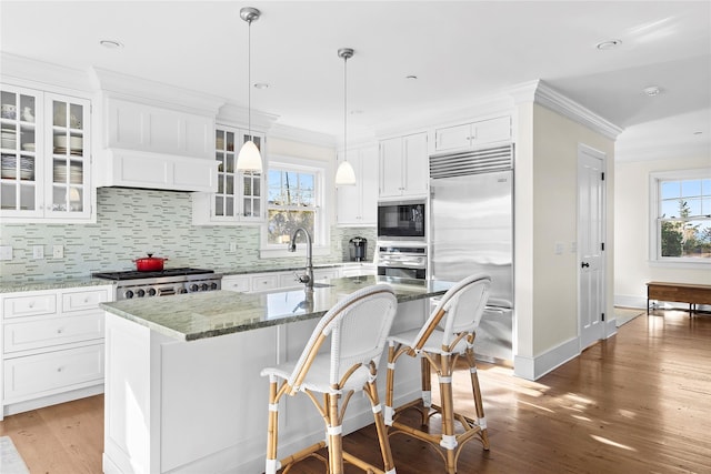 kitchen featuring white cabinetry, built in appliances, dark stone counters, a breakfast bar area, and a center island with sink