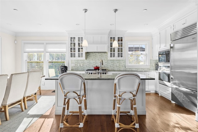 kitchen with built in appliances, a center island with sink, white cabinets, and hanging light fixtures