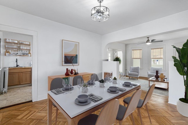 dining space with radiator, sink, light parquet flooring, and ceiling fan with notable chandelier