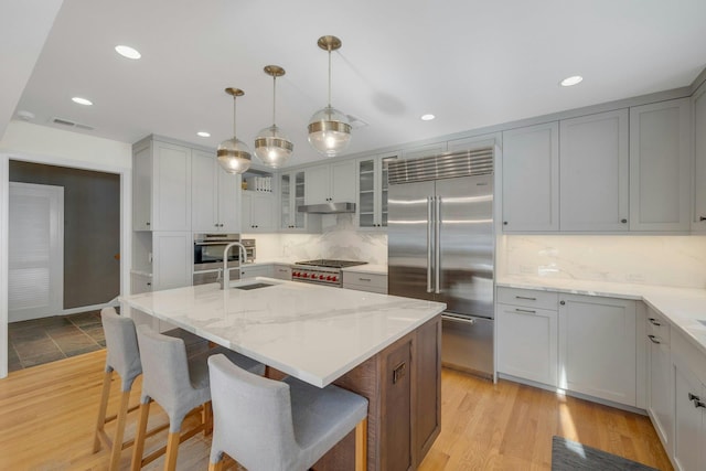 kitchen with sink, a kitchen island with sink, hanging light fixtures, stainless steel appliances, and light stone counters