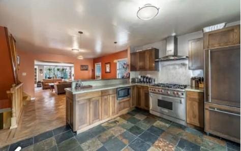 kitchen with kitchen peninsula, backsplash, wall chimney exhaust hood, high end appliances, and decorative light fixtures