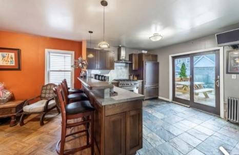 kitchen with appliances with stainless steel finishes, radiator, a healthy amount of sunlight, wall chimney range hood, and hanging light fixtures