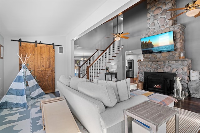 living room featuring hardwood / wood-style flooring, ceiling fan with notable chandelier, high vaulted ceiling, a stone fireplace, and a barn door