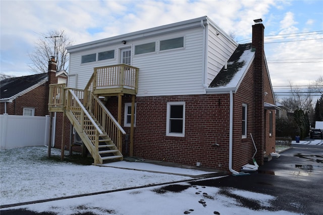 snow covered back of property with brick siding, fence, a chimney, and stairs