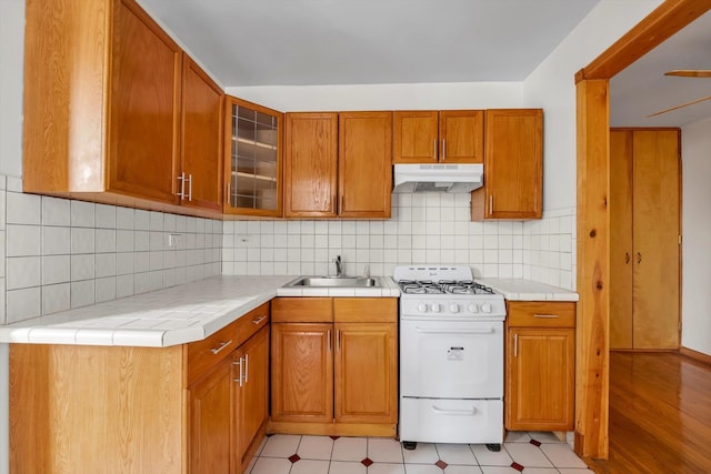 kitchen featuring glass insert cabinets, brown cabinets, white gas stove, under cabinet range hood, and a sink