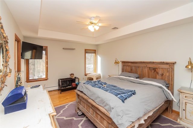 bedroom with ceiling fan, a raised ceiling, light wood-type flooring, and a baseboard heating unit