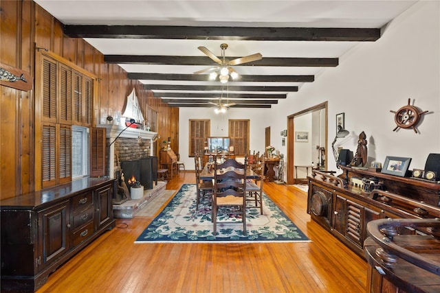 dining room featuring ceiling fan, beamed ceiling, wood walls, a fireplace, and light wood-type flooring