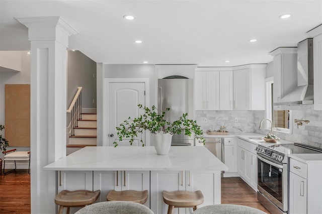 kitchen with wall chimney range hood, sink, white cabinetry, stainless steel appliances, and a kitchen island