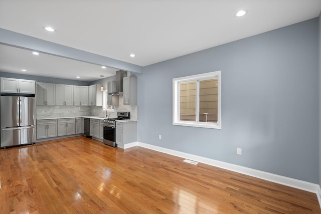 kitchen with gray cabinetry, stainless steel appliances, wall chimney range hood, tasteful backsplash, and light hardwood / wood-style floors