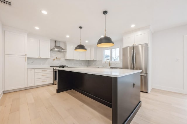 kitchen with wall chimney range hood, white cabinets, light hardwood / wood-style flooring, a kitchen island, and appliances with stainless steel finishes