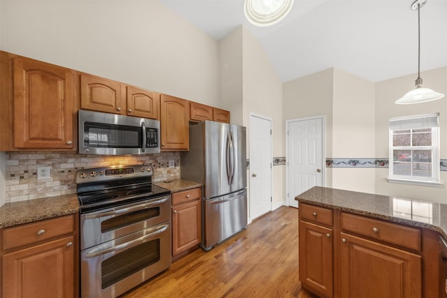 kitchen featuring dark stone countertops, appliances with stainless steel finishes, hanging light fixtures, light hardwood / wood-style flooring, and tasteful backsplash