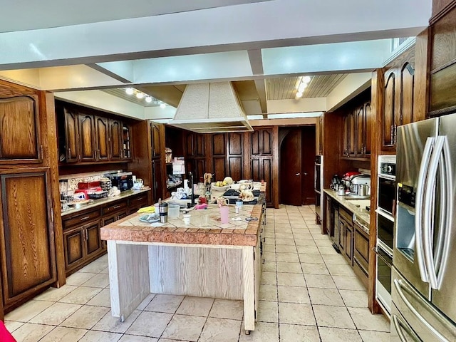 kitchen with custom exhaust hood, a kitchen island with sink, appliances with stainless steel finishes, light tile patterned flooring, and dark brown cabinetry