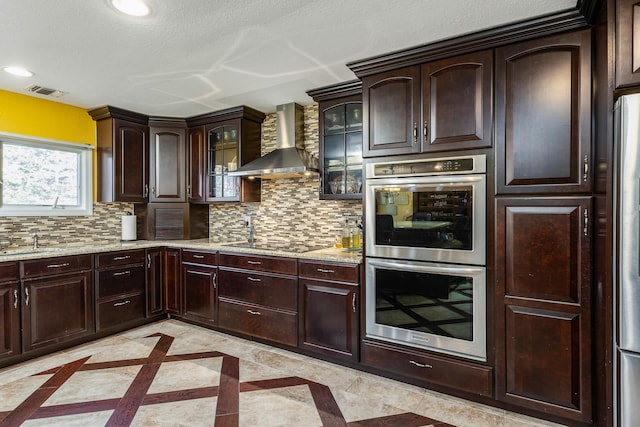 kitchen with stainless steel double oven, wall chimney range hood, light stone counters, decorative backsplash, and black electric stovetop