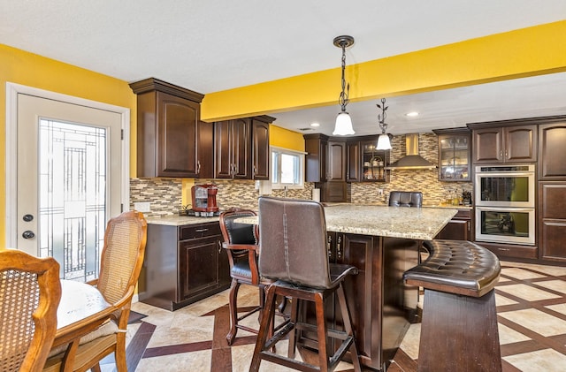 kitchen with dark brown cabinetry, wall chimney exhaust hood, double oven, backsplash, and a kitchen island