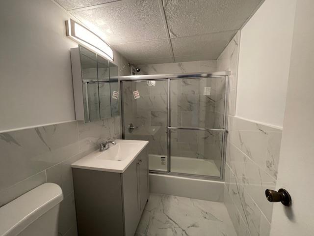 full bathroom featuring toilet, vanity, tile walls, combined bath / shower with glass door, and a paneled ceiling