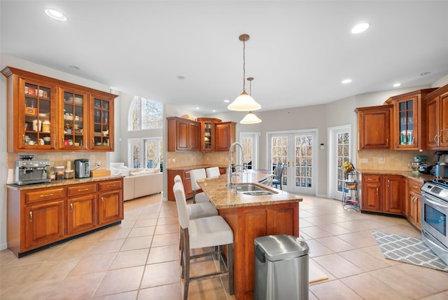 kitchen with sink, light stone counters, an island with sink, stainless steel range, and pendant lighting