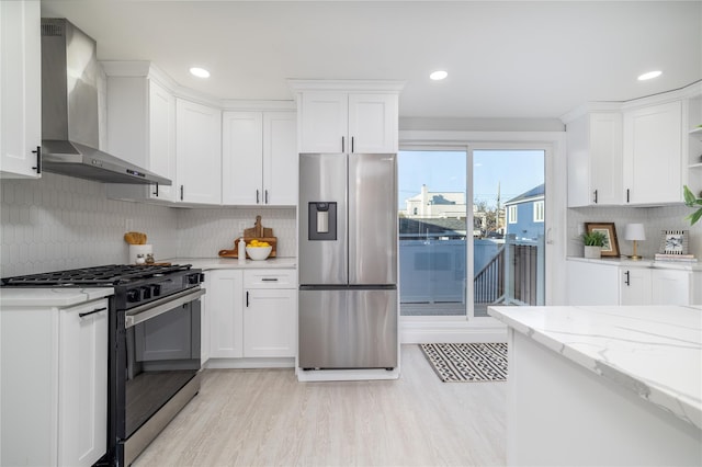 kitchen featuring wall chimney range hood, light stone countertops, tasteful backsplash, white cabinetry, and stainless steel appliances