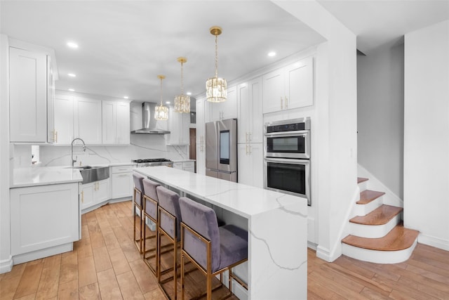 kitchen with appliances with stainless steel finishes, white cabinets, a sink, a kitchen island, and wall chimney exhaust hood