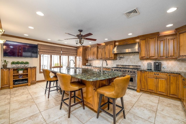 kitchen featuring wall chimney range hood, dark stone countertops, a kitchen island with sink, and stainless steel stove