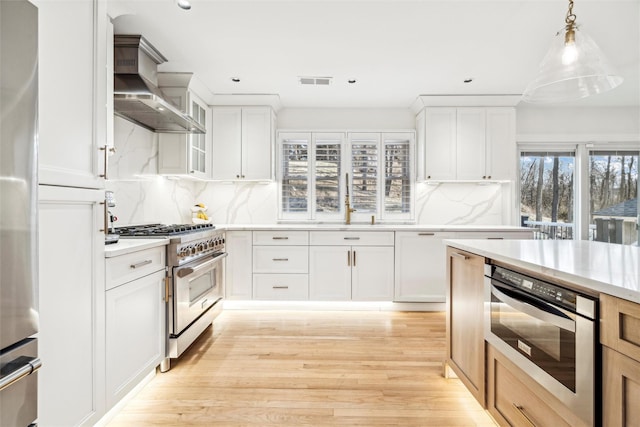 kitchen featuring stainless steel appliances, white cabinetry, hanging light fixtures, and wall chimney range hood