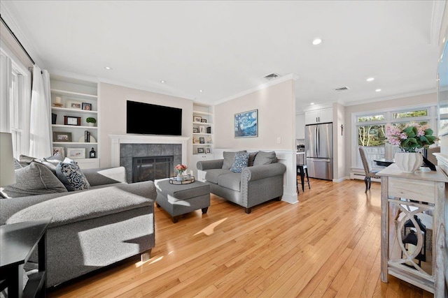 living room with a tiled fireplace, crown molding, built in features, and light hardwood / wood-style floors