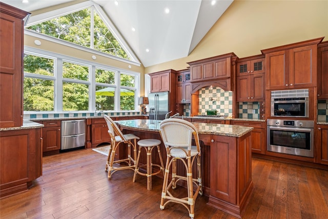 kitchen with dark wood-type flooring, appliances with stainless steel finishes, backsplash, light stone counters, and a kitchen island