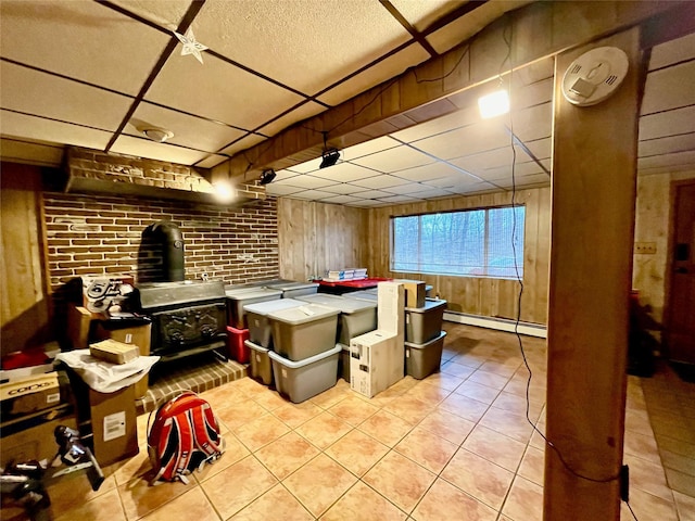 interior space featuring a wood stove, a drop ceiling, baseboard heating, brick wall, and wood walls