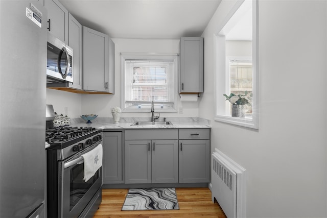 kitchen featuring gray cabinetry, sink, radiator heating unit, stainless steel appliances, and light stone counters