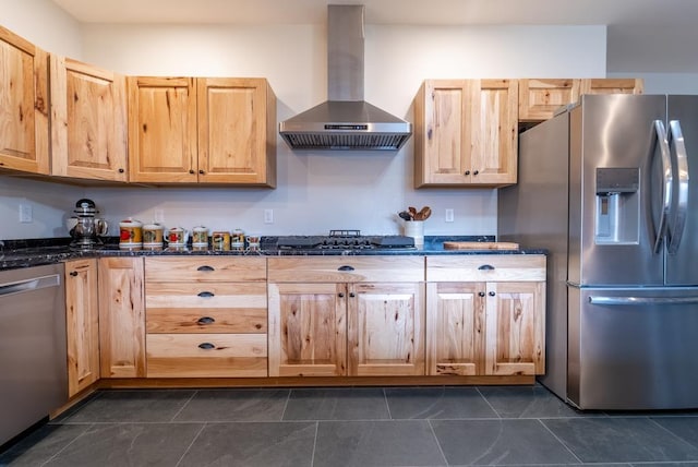 kitchen with light brown cabinetry, stainless steel appliances, dark stone countertops, and wall chimney range hood