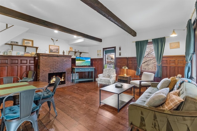 living area with a wainscoted wall, dark wood-type flooring, beamed ceiling, and a brick fireplace