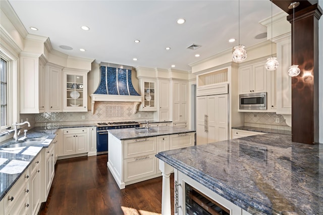 kitchen with sink, hanging light fixtures, built in appliances, dark stone counters, and custom range hood