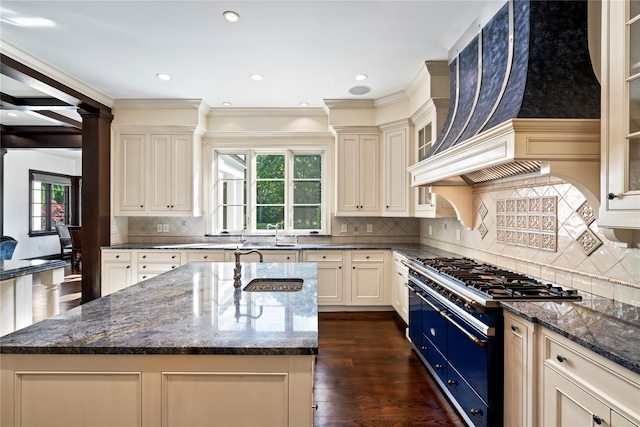 kitchen featuring gas range, dark wood-type flooring, cream cabinets, dark stone counters, and a kitchen island with sink