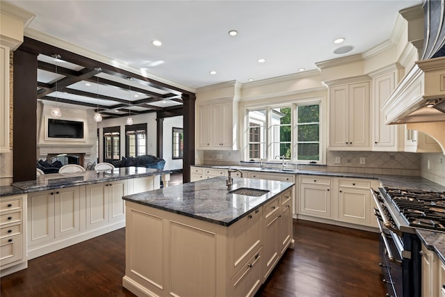 kitchen featuring dark stone counters, sink, beam ceiling, cream cabinets, and a center island with sink