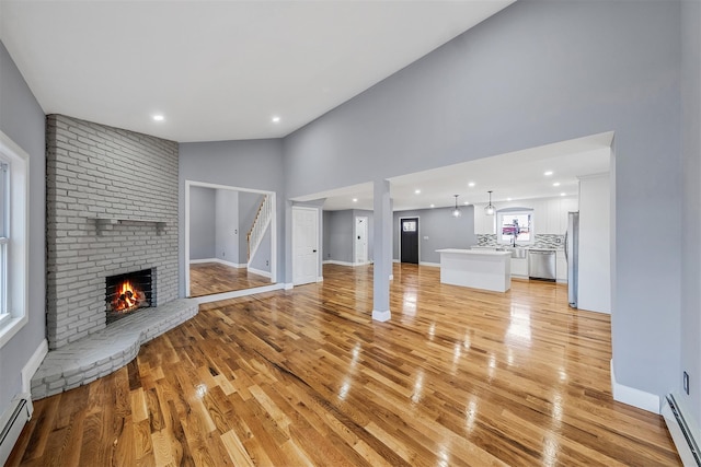 unfurnished living room featuring light hardwood / wood-style flooring, high vaulted ceiling, a baseboard radiator, and a brick fireplace