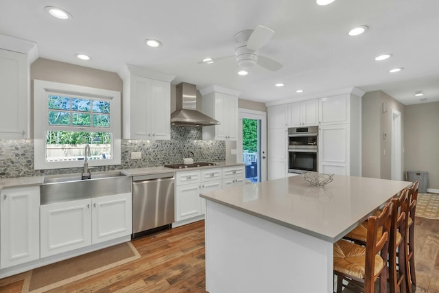 kitchen with a center island, white cabinets, wall chimney exhaust hood, appliances with stainless steel finishes, and light hardwood / wood-style floors