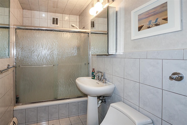 bathroom featuring sink, a baseboard heating unit, toilet, a shower with door, and tile walls