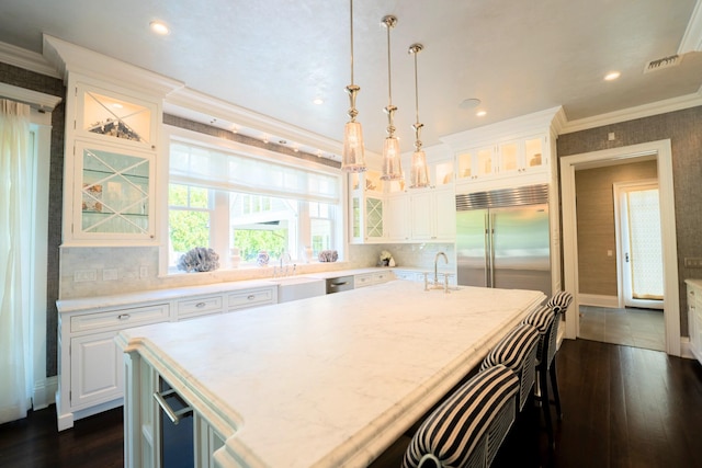 kitchen with built in refrigerator, pendant lighting, white cabinets, and an island with sink