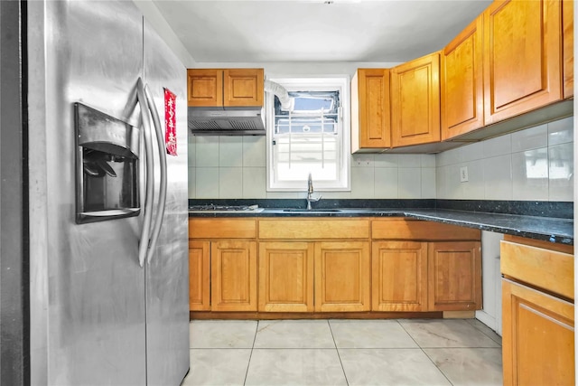 kitchen with sink, dark stone countertops, light tile patterned floors, range hood, and stainless steel refrigerator with ice dispenser