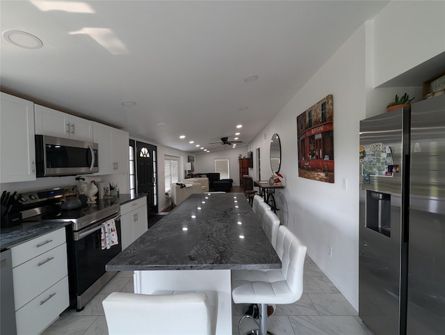 kitchen with dark stone counters, white cabinets, ceiling fan, appliances with stainless steel finishes, and a breakfast bar area