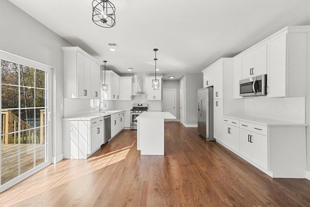 kitchen featuring appliances with stainless steel finishes, wall chimney exhaust hood, a kitchen island, pendant lighting, and white cabinetry