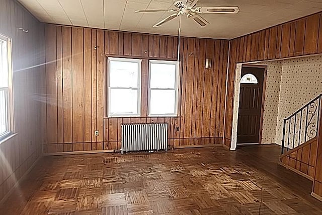 empty room with dark parquet floors, radiator, wood walls, and ceiling fan