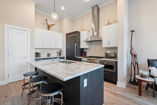 kitchen with white cabinetry, a kitchen island with sink, pendant lighting, wall chimney exhaust hood, and sink