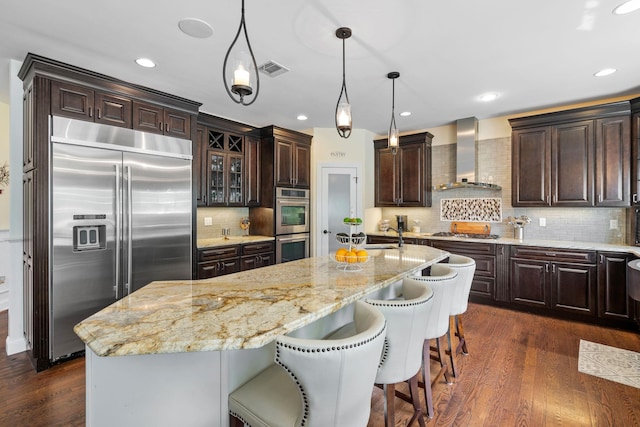 kitchen with dark brown cabinets, a kitchen island with sink, wall chimney range hood, and stainless steel appliances