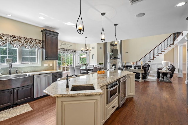 kitchen featuring hanging light fixtures, dark brown cabinetry, an island with sink, stainless steel appliances, and light stone counters