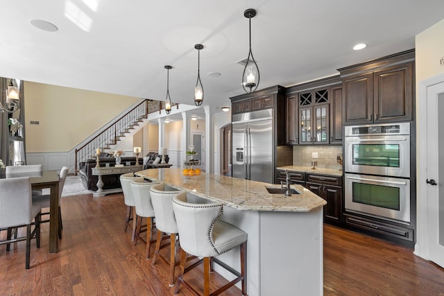 kitchen with dark brown cabinets, hanging light fixtures, appliances with stainless steel finishes, an island with sink, and decorative columns