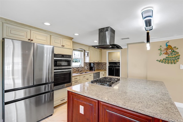 kitchen with island exhaust hood, tasteful backsplash, cream cabinets, appliances with stainless steel finishes, and a sink