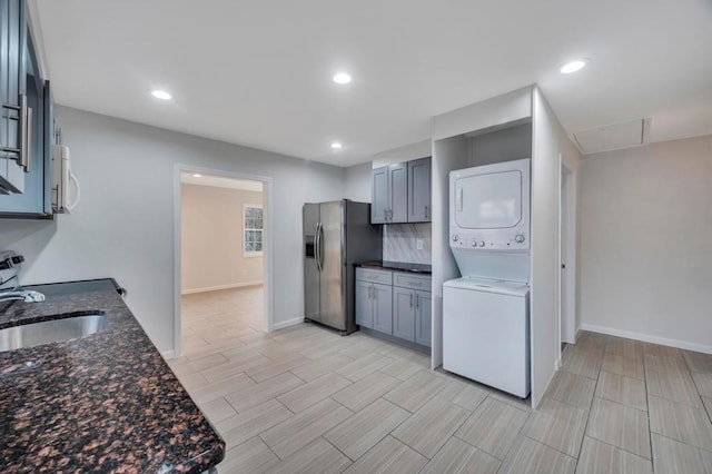 kitchen with sink, stacked washer and clothes dryer, dark stone counters, stainless steel refrigerator with ice dispenser, and gray cabinetry