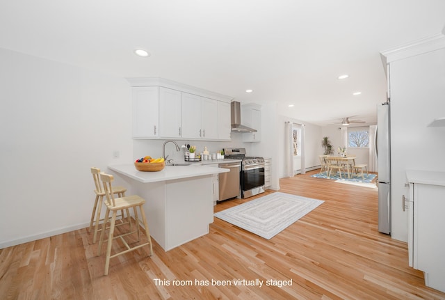 kitchen featuring kitchen peninsula, stainless steel appliances, wall chimney range hood, a kitchen bar, and white cabinetry